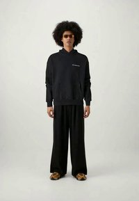 Νέος άνθρωπος με αφρικανικό χτένισμα, φορά καφέ γυαλιά ηλίου, μαύρη κουκούλα με κείμενο, φαρδιά μαύρα παντελόνια και καφέ-πορτοκαλί αθλητικά παπούτσια, ενώ στέκεται.