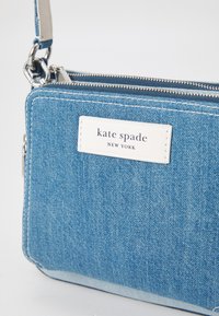 Denim crossbody bag with a blue exterior, white logo patch, silver hardware, a zipper closure, and a detachable strap.