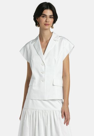Žena oblečená v bielom bezrukávovom blejzeri s tromi gombíkmi a klopovými vreckami, v kombinácii s bielou nariasenou sukňou na jednoduchom pozadí.