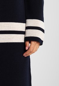 Marinblå stickad tröja med krämfärgade och marinblå randiga muddar. Mjuk struktur, avslappnad passform. Har långa ärmar och en rak silhuett.