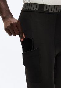 Χέρι που βάζει ένα μαύρο smartphone στην πλαϊνή τσέπη μαύρων κολάν Puma με άσπρο πουκάμισο περασμένο μέσα.