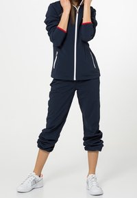 Marinblå jacka med röda detaljer och vit dragkedja, ihop med korta marinblå byxor. Båda är tillverkade av lättviktsmaterial; vita sneakers fulländar outfiten.