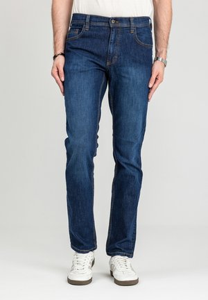 Mężczyzna ubrany w ciemnoniebieskie dżinsy o prostych nogawkach i białe sneakersy, stojący na tle jednolitego jasnego tła.