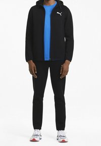 Sudadera negra con cremallera y capucha, con un logo blanco. Combinada con una camisa azul y pantalones negros, complementada por zapatillas deportivas coloridas.