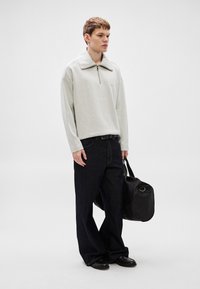 Ανοιχτό γκρι πουλόβερ με ψηλό γιακά και φερμουάρ τετάρτου, συνδυασμένο με σκούρα μπλε τζιν παντελόνια φαρδιάς γραμμής και μαύρη τσάντα τύπου duffle. Ομαλή υφή, casual εφαρμογή.