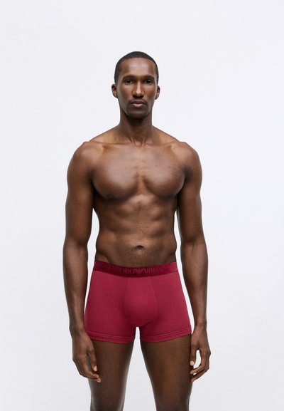 Burgundske bokserice s gladko teksturo in elastičnim pasom, ki nosi logo "Emporio Armani" v temno rdeči barvi. Model stoji obrnjeno naprej.