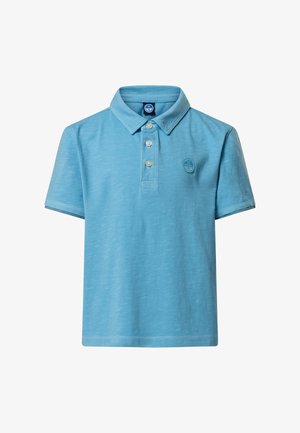 Jeune garçon aux cheveux châtain clair courts portant un polo bleu clair et un pantalon blanc, debout devant un fond uni gris clair.