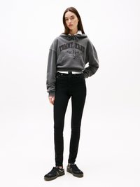 Kvinna i en grå Tommy Jeans hoodie, svarta tajta jeans och svarta sneakers, står med en hand i fickan mot en vit bakgrund.