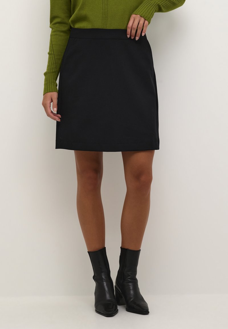Femme portant une jupe noire jusqu'aux genoux, un pull vert côtelé et des bottines noires, se tenant devant un fond clair uni.