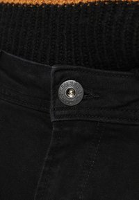 Musta farkkuhousut, joissa on nappisuljenta, näkyvät ompelusaumat ja musta-oranssilla kuvioitu neulottu vyötärönauha.