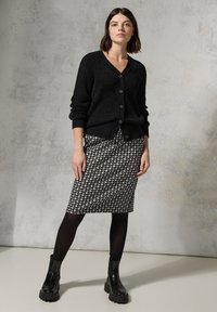 Schwarze, strukturierte Strickjacke mit Knöpfen, kombiniert mit einem Bleistiftrock im Hahnentrittmuster. Das Model trägt schwarze Knöchelstiefel und Strumpfhosen.