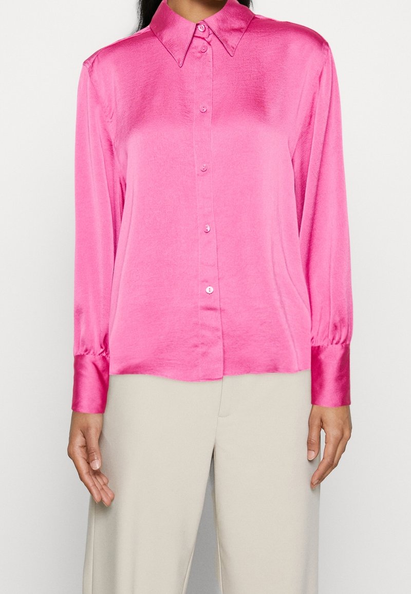 Femme portant un chemisier long à manches longues en satin rose vif avec col pointu et un pantalon beige taille haute, sur un fond uni.