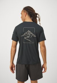 Camiseta de manga corta negra hecha de tejido texturizado, con un gráfico de montaña y río en gris en la parte posterior. Lleva puesta con pantalones cortos oscuros.