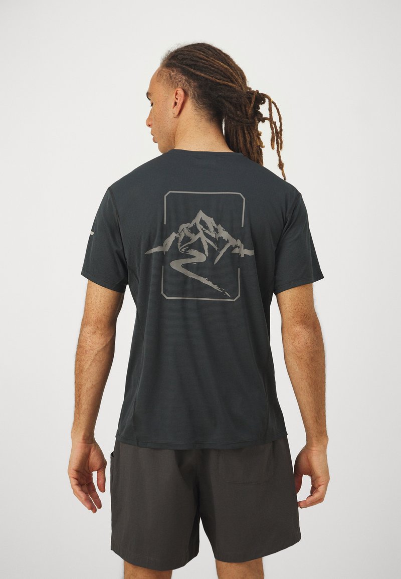 Camiseta de manga corta negra hecha de tejido texturizado, con un gráfico de montaña y río en gris en la parte posterior. Lleva puesta con pantalones cortos oscuros.