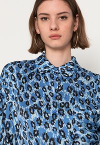 Camisa de estampa de leopardo azul com colarinho, apresentando padrões em preto e azul claro, fechamento com botões e um bolso do lado esquerdo.
