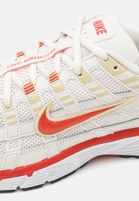 Białe siatkowe buty sportowe z czerwonymi i złotymi akcentami, z wyraźnym czerwonym logo Nike, fakturowanymi sznurówkami oraz białą podeszwą środkową.