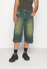Pantalones cortos de mezclilla en un color verde desvaído, con un ajuste holgado, dobladillo vuelto y bolsillos delanteros, combinados con zapatillas negras y calcetines cortos.