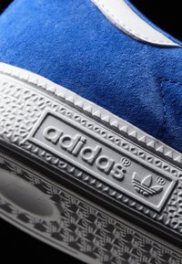 Blå mockasneaker med vita detaljer, försedd med en texturerad gummisula och en präglad logotypplåt. Designen betonar rena linjer och detaljer.