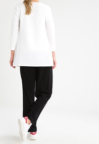 Langärmlige weiße Tunika, die mit schwarzen Hosen kombiniert ist. Die Tunika hat eine strukturierte Oberfläche; die Hosen sind locker geschnitten. Weiße Sneaker mit rosa Akzenten.