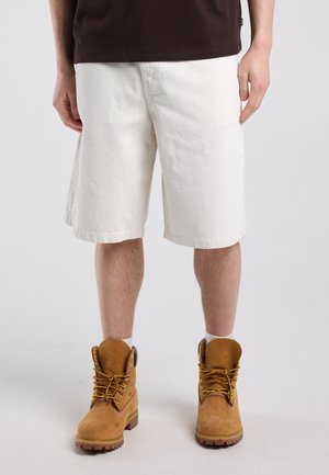 Persoon die off-white knielange shorts draagt, bruine werkschoenen met gele veters en een donkerbruine blouse, staand op een witte achtergrond.