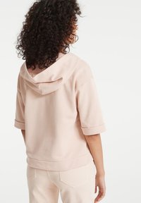 Felpa rosa a maniche corte con una texture morbida, munita di un cappuccio ampio e polsini arrotolati. Il materiale sembra leggero e informale.