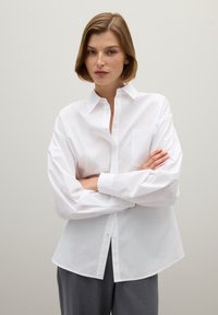 White button-up shirt made of smooth fabric, featuring a pocket on the left side, a classic collar, and long, slightly puffed sleeves.