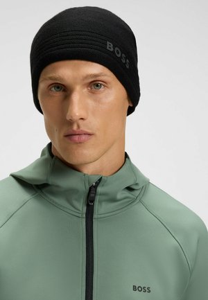 Black knitted beanie with textured design and "BOSS" logo in gray. Worn with a green zip-up jacket featuring a high collar and zipper detail.