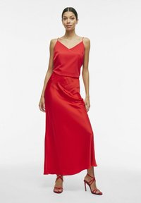 Rode satijnen maxi-jurk met dunne bandjes en een aansluitende bodice, vloeiende rok en een soepele textuur. Gecombineerd met rode strappy hakken.