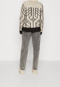 Stickad tröja i beige med svarta geometriska mönster, i kombination med grå denimjeans och ljusa stadiga sneakers.