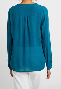 Blouse à manches longues couleur sarcelle avec un col rond, un détail plissé dans le dos et des poignets boutonnés. Confectionnée en tissu léger et texturé.