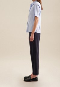 Ljusblå kortärmad skjorta, sidovy; i kombination med marinblå byxor och svarta loafers. Slät tyg, avslappnad passform, med knapp-up krage.
