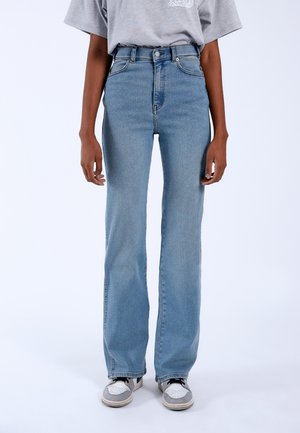 Person iført lyseblå jeans med højtaljet, lige ben, grå t-shirt og hvide-grå sneakers, stående mod en ensfarvet baggrund.