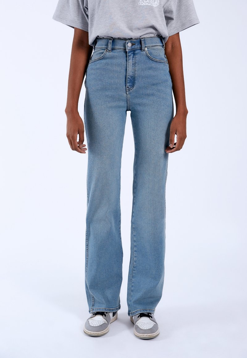 Personne portant un jean droit taille haute bleu clair, un t-shirt gris et des baskets blanc-gris, debout devant un fond uni.