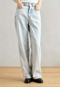 Jean en denim bleu clair avec une coupe ample, taille haute, poches avant et détails de décoloration subtils. Portés avec des chaussures noires sur un sol en bois.