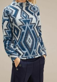 Neulepusero geometrisilla kuvioilla sinisen, harmaan ja valkoisen sävyissä. Varustettu korkealla kauluksella ja pitkillä hihoilla. Yhdistetty tummansinisiin housuihin.