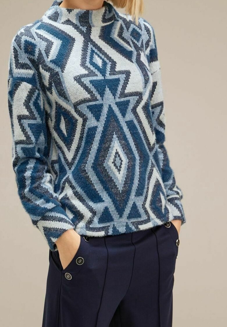 Neulepusero geometrisilla kuvioilla sinisen, harmaan ja valkoisen sävyissä. Varustettu korkealla kauluksella ja pitkillä hihoilla. Yhdistetty tummansinisiin housuihin.