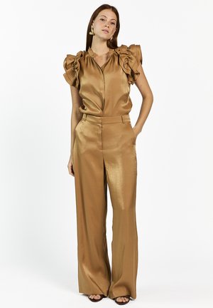 Vrouw draagt een glanzende bronzen blouse met ruches aan de mouwen en bijpassende wijde broekspijpen, staat met één hand in de zak.