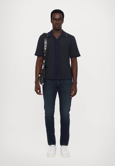 Kortermet, marineblå ribbestrikket skjorte med tynn krage, kombinert med mørke jeans og hvite sneakers. Modellen bærer en svart veske.