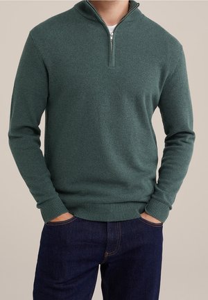 Mand iført en tekstureret mørkegrøn kvartlyn lås sweater og mørkeblå jeans med hænderne i lommerne mod en ensfarvet baggrund.