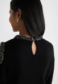 Μαύρο πουλόβερ με κολάρο διακοσμημένο με χάντρες, άνοιγμα στον αυχένα και υφή πλεγμένου υφάσματος. Περιλαμβάνει κουμπί στο λαιμό.