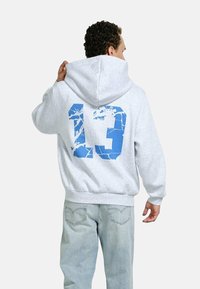 Man wearing light gray hoodie with large cracked blue number 13 on the back, paired with light blue jeans, facing away.