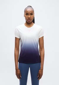 Γυναίκα με μαλλιά δεμένα πίσω, που φοράει μπλουζάκι με βαθμιαία μετάβαση από λευκό σε σκούρο μπλε με κοντά μανίκια και μπλε κολάν, στέκεται μπροστά σε απλό φωτεινό φόντο.