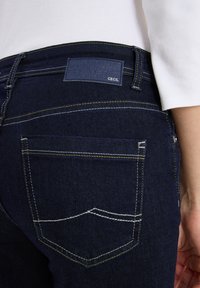 Pantalones vaqueros de mezclilla azul oscuro con costuras amarillas y blancas, parche de marca negro que dice "Cecil" en la cintura trasera, usados con una blusa blanca.
