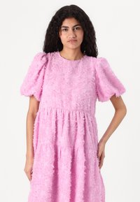 Vestido rosa em camadas com um design de franjas texturizadas, mangas curtas bufantes e decote arredondado. O tecido parece suave e leve.