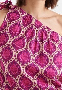 Top na jedno ramię z zmarszczonym wykończeniem, w kolorze kremowym, z jaskrawym różowym i fioletowym wzorem paisley. Zaprojektowany z detalem w postaci związanej ramiączka.