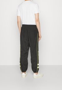 Svarta sportbyxor med elastiska avslut, med gula ränder och "LACOSTE"-logotyp på sidan. Tillverkade av lätt material.