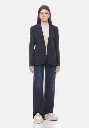 Donna in piedi, indossa un blazer blu scuro, una maglia beige, jeans larghi a gamba larga di colore scuro e sneakers beige con plateau su uno sfondo bianco semplice.