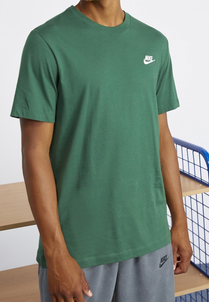 Muž v zeleném tričku Nike a šedých šortkách Nike stojí u dřevěných regálů a modrého kovového rámu.