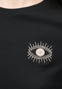 Μαύρο ύφασμα με λευκό κεντητό σχέδιο ματιού που περιλαμβάνει βλεφαρίδες στο πάνω μέρος του στήθους ενός πουκάμισου ή τοπ.