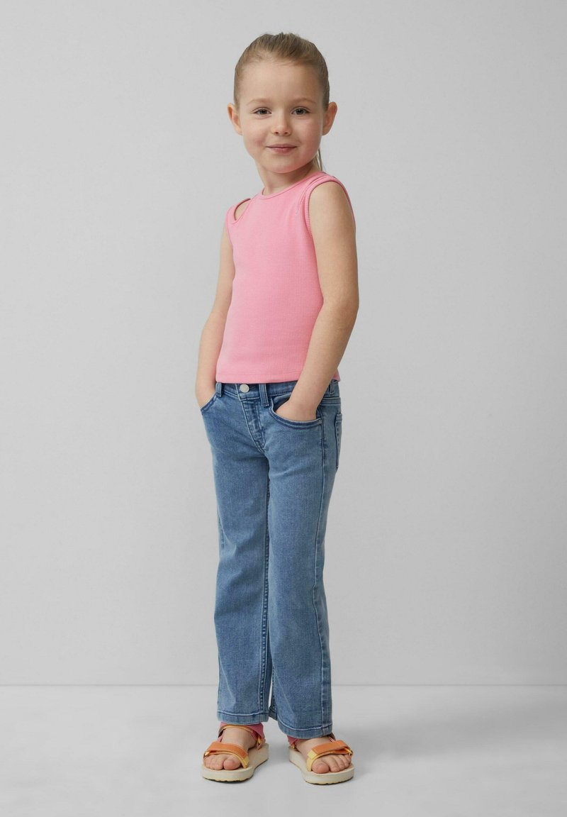 Roze mouwloze ribbed top, lichtblauwe jeans met een lichte flare, en oranje sandalen met bicolor bandjes, handen in de zakken, neutrale achtergrond.